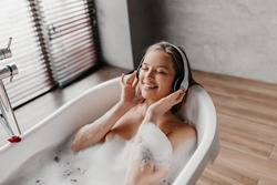 Charming woman lying in hot bubbly bath and listening to music in wireless headphones, free space. Pretty young lady enjoying favorite playlist on domestic spa day