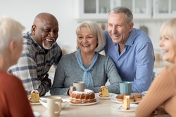 Cheerful senior people of different nationalities sitting at kitchen, drinking tea and eating cake together, having conversation and laughing, chilling together at nursing home