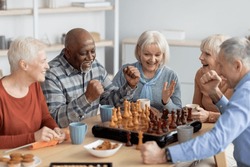 Emotional multiethnic group of senior people playing chess, happy elderly men and women in casual outfits sitting around table with table games, chatting, having fun at sanatorium