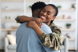Closeup portrait of cheerful black female soldier lady in camouflage uniform hugging her boyfriend, home interior. Military wife came back from army, embracing her loving husband, finally together