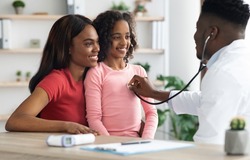 Smiling african american girl visiting doctor at clinic, sitting with her mother in front of pediatrician, black man in uniform doc listening to child patient breath with stethoscope, panorama