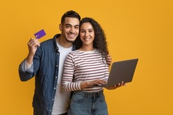 Online Shopping Concept. Happy Arabic Couple Using Credit Card And Laptop While Standing Over Yellow Background In Studio, Cheerful Middle Eastern Man And Woman Enjoying Purchasing In Internet
