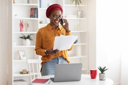 Young businesswoman cheerful african american lady with red turban on her head working at office, standing by workdesk with laptop, holding documents, having phone conversation, copy space