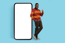 Online Promo. Joyful Black Man Standing Near Big Blank Smartphone And Showing Thumb Up, Cheerful African American Male Leaning At Mobile Phone With White Screen For Mockup, Full Length Shot