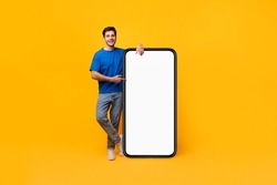 Full Body Length Of Happy Casual Guy Pointing Finger At Big Blank Smartphone Display Leaning On Huge White Empty Cell Phone Screen Standing On Yellow Studio Background Mockup. Mobile App Advertisement