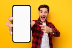 Great Offer. Excited surprised guy holding and pointing finger at big giant cell phone with white screen in hand, promoting application or website, advertising product or service, yellow studio wall