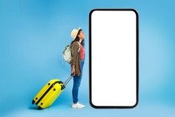 Young black female traveler with suitcase and backpack looking at big smartphone with empty screen, booking hotel or vacation online, offering mockup for online travel agency website, blue background
