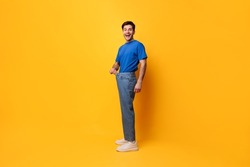 Successful Weight Loss. Happy Excited Slim Young Guy Pulling Old Large Loose Jeans Achieved Slimming Weightloss Goal Standing Posing Isolated On Yellow Studio Wall, Full Body Length, Free Copy Space