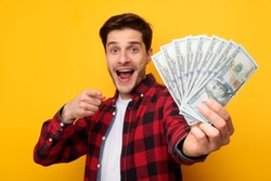 Portrait Of Excited Man Holding Fan Of Money Cash In Hands Pointing Finger Showing It To Camera On Yellow Orange Background. Guy Posing With Banknotes. Financial Success And Currency Concept. Big Luck