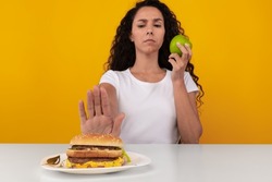 No Junk Food. Serious Lady Refusing To Eat Unhealthy Burger Choosing Apple, Showing Stop Sign Gesture With Palm Sitting At Table, Selective Focus On Cheeseburger And Hand. Healthy Eating, Diet Concept