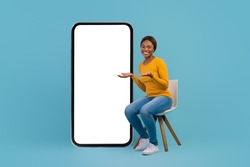 Cheerful Black Woman Sitting On Chair Near Big Blank Smartphone, Happy Young African American Lady Recommending New Mobile App Or Website, Posing On Blue Studio Background, Collage, Mockup