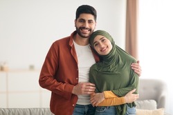 Loving Middle Eastern Couple Embracing Posing Standing At Home. Muslim Husband Hugging His Wife With Hijab Smiling To Camera. Love And Romance, Happy Relationship And Marriage Concept