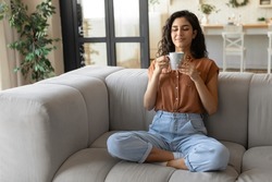 Peaceful young brunette woman sitting on couch with closed eyes, drinking hot coffee at home, copy space. Full length of millennial female enjoying lazy morning with warm drink in living room