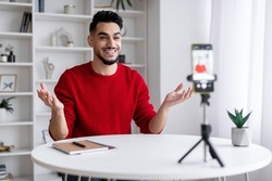 Young Arab Male Blogger Recording Video At Home With Smartphone On Tripod, Smiling Middle Eastern Guy Sitting At Desk And Making Content For Blog, Talking And Gesturing At Phone Camera, Free Space