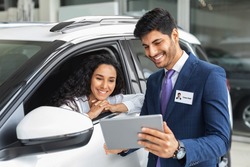 Pretty long-haired brunette lady sitting inside brand new comfy white car, having conversation with cheerful sales assistant, looking at digital tablet in dealer hands, checking auto features