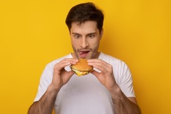 Guilty Pleasure Concept. Portrait of hungry funny young man holding burger in hands and eating unhealthy fastfood, excited starving guy wants sandwish licking his lips, yellow orange studio wall