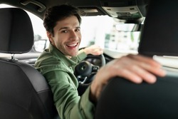 Cheerful young man driving new automobile after purchase, sitting in driver's seat of car at dealership. Happy millennial guy looking back and smiling, buying modern auto at showroom