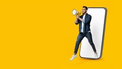 Handsome young arab man jumping out of huge smartphone with white empty screen, holding megaphone over yellow studio background, making important announcement, panorama with copy space