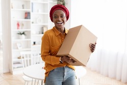 Happy young african american muslim woman in casual holding box with something and smiling, home interior, making order on Internet with delivery, copy space. Delivery service, shipment concept