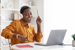 Joyful short-haired millennial black lady having break while working at office, watching video on laptop, using wireless headset, holding pens and playing imaginary drums, relaxing, copy space