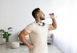 Sporty young Arab man with headphones drinking water after his workout at home. Fit Eastern guy staying hydrated during domestic sports training. Healthy lifestyle, wellness concept