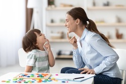 Speech training for kids. Professional woman specialist training with little boy at cabinet, teaching him right articulation exercises, side view