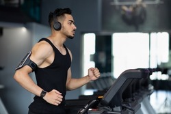 Muscular Arab Male Athlete Wearing Wireless Headphones Running On Treadmill At Gym, Sporty Millennial Middle Eastern Guy Listening Music During Workout, Enjoying Cardio Training, Copy Space