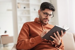 Portrait of focused young Arabic male trying to read paper book, squinting to see more clearly, wearing glasses, having difficulties seeing text because of vision problems, sitting on couch