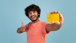 Cheerful handsome young indian man holding yellow credit card and showing thumb up, pleased client demonstrating bank product over blue studio background, panorama. Finance, loan, savings concept