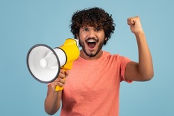 Motivated curly eastern millennial guy in casual shouting with loudspeaker and gesturing, making announcement over blue studio background, sharing his emotions, chanting slogans, copy space