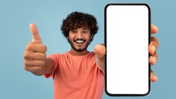 Handsome indian millennial guy showing brand new smartphone with empty white screen and thumb up, smiling at camera over blue studio background, mockup, mobile application concept, panorama