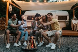Millennial diverse friends sitting near bonfire with lit sparklers, having fun, celebrating something together on camping trip, traveling in motorhome, enjoying friendly conversation outdoors