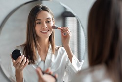Attractive Young Woman Doing Daily Makeup While Standing Near Mirror In Bathroom, Happy Beautiful Female Putting Blush On Cheeks While Getting Ready At Home, Selective Focus On Reflection