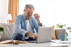 Omg, Wow. Portrait of surprised mature man sitting at desk on couch using laptop, taking off glasses looking at pc screen in amazement. Emotional male reading breaking news online in home office