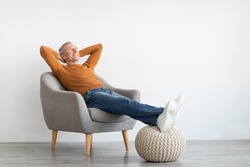 Smiling calm mature man relaxing sitting on armchair, resting feet on knitted pouf, free copy space. Happy adult spending weekend, leaning back holding hands behind head, isolated on white studio wall