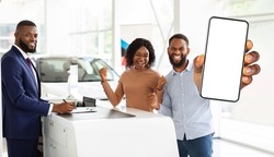 Car Insurance. Happy Black Spouses Showing Blank Smartphone At Camera While Standing Next To Salesman In Auto Showroom, Signing Contract With Friendly Manager After Purchasing New Automobile, Mockup