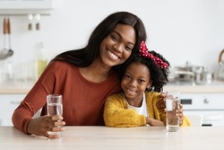 Happy African American Family Mother And Daughter Holding Glasses With Water While Sitting Together At Table In Kitchen, Smiling Black Mom And Female Child Enjoying Healthy Drink, Free Space