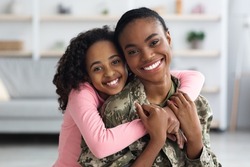 Portrait of cheerful pretty curly black teen girl hugging her mom soldier from behind and smiling at camera, young black woman in military uniform return home from army, cuddling with her daughter