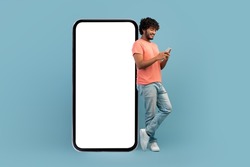 Cool curly indian guy in casual outfit leaning on huge smartphone with white blank screen, using cell phone and smiling, blue studio background, mockup, panorama with copy space