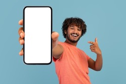 Cool handsome curly young indian guy in stylish pink t-shirt showing brand new cell phone with white empty screen and gesturing, sharing exciting online deal, blue background, mockup