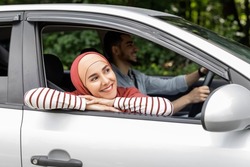 Smiling millennial arab female in hijab ride in car looks out the open window outdoor, enjoy journey, man drives car. Travel in summer, weekends on road, holidays with family after covid-19 pandemic