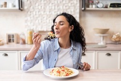 Young latin woman eating delicious pasta, enjoying tasty homemade lunch with closed eyes while sitting at table in light kitchen interior, free space
