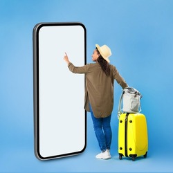 African Woman Traveler Using Huge Smartphone Touching Empty White Mobile Touchscreen Browsing Internet Booking Flight Tickets Standing Near Cellphone With Suitcase On Blue Background. Square