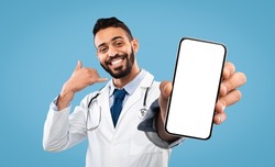 Cheerful Arab Doctor Male Gesturing Call Me, Holding Cell Phone With White Empty Screen, Posing On Blue Studio Background, Smiling To Camera. Medical Consultation By Phone Concept, Mockup