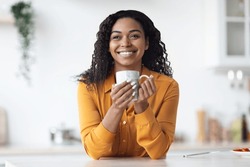 Closeup of happy pretty long-haired young black woman in yellow shirt sitting at kitchen table with cup of herbal tea, looking at copy space and smiling, enjoying morning coffee at home