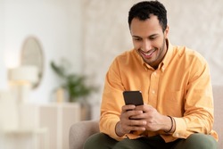 People And Gadgets. Cheerful Middle Eastern Man Using Cellphone Playing Mobile Game Online, Browsing Internet And Texting Sms Sitting On Couch In Living Room Indoors. Cellular Communication Concept