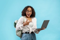 Big sale, great luck concept. Emotional young black woman with backpack pointing at laptop screen in excitement over blue studio background. Great online educational course offer