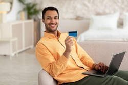 Easy Online Payments. Smiling Arabic Man Using Laptop And Holding Showing Credit Card In Hand, Happy Relaxed Guy Sitting On Comfortable Couch, Shopping In Internet Transferring Money Looking At Camera