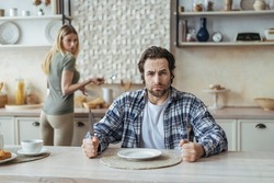 Angry hungry dissatisfied millennial caucasian man with stubble ignoring woman and wait for food in kitchen interior. Quarrel, family problems, stress, social issues and pressure due covid-19 pandemic