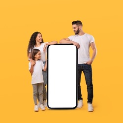 Middle Eastern Family Leaning On Huge Smartphone With Blank Screen Advertising Mobile App On Yellow Background. Parents And Daughter Standing Near Phone Recommending Mobile Offer. Mockup, Square